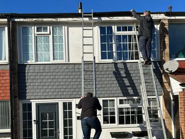 Two Homeguard roofers on ladders fitting new fascias, guttering and grey cladding to the front of a terraced house