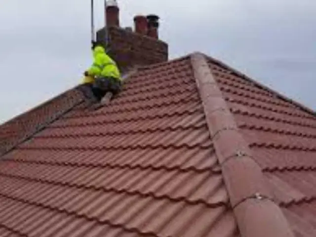 A roofer in a high-visibility jacket at the ridge of a red tiled roof beside a chimney stack, on a grey day