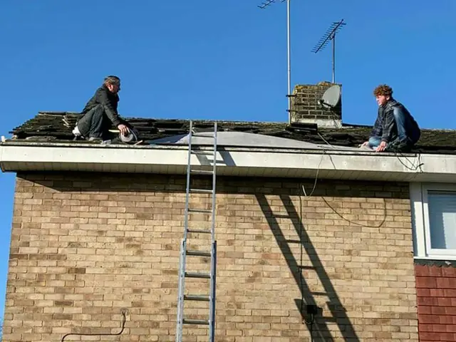 Two roofers kneeling on a tiled roof above a brick wall, repairing the tiles under a clear blue sky
